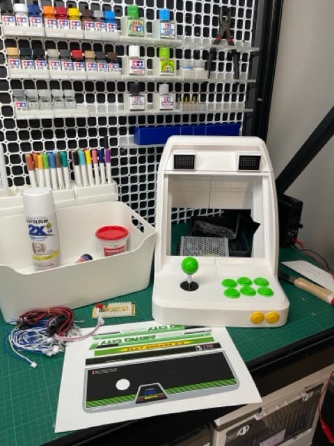 A printed white bartop arcade cabinet with a joystick and green buttons, on a workbench beside a rack of Tamiya paints and its printed artwork panel