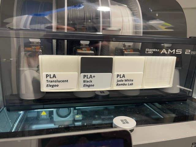 Printed two-colour labels clipped to the front of a Bambu Lab AMS 2 Pro, reading PLA Translucent, PLA+ Black and PLA Jade White